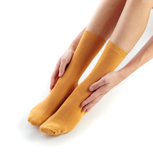 A pair of mustard yellow crew socks worn on a person's legs, with hands adjusting them against a clean white background.
