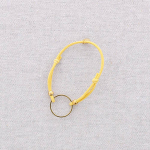 A delicate yellow cord bracelet with a small gold ring charm, displayed on a textured light gray fabric background.