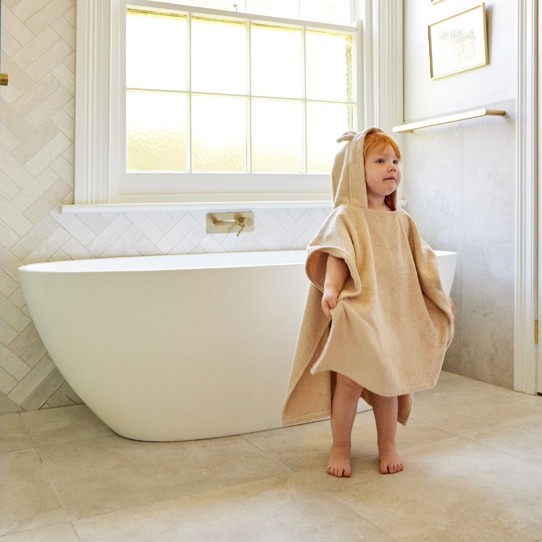 Child wearing a beige hooded towel standing in a bathroom with a freestanding bathtub.
