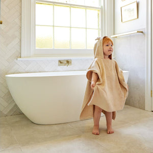 Child wearing a beige hooded towel standing in a bathroom with a freestanding bathtub.