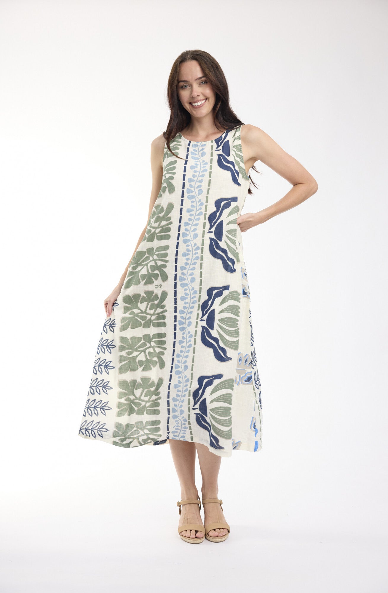 A woman wearing a sleeveless midi dress with a botanical leaf pattern in green, navy, and blue on an off-white background, standing on a sunlit patio with plants and the ocean in the background.