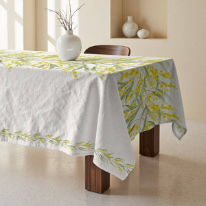 A white cotton tablecloth with a yellow and green floral pattern, draped over a wooden dining table in a warm, minimalist room.