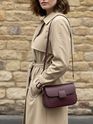 Woman wearing a beige trench coat with a burgundy handbag against a stone wall.