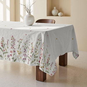 A white tablecloth with a delicate multicolor floral print draped over a wooden dining table in a warm, minimalist room.