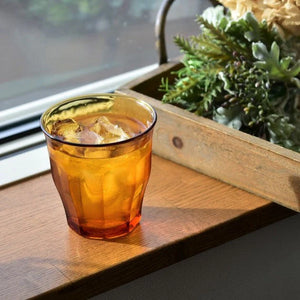 Amber-colored glass with a drink and ice cubes on a wooden surface with a plant in the background
