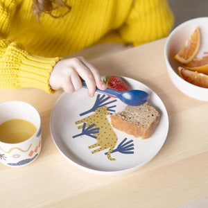 Child's hand holding a spoon over a plate with a leopard design, surrounded by food on a wooden table.