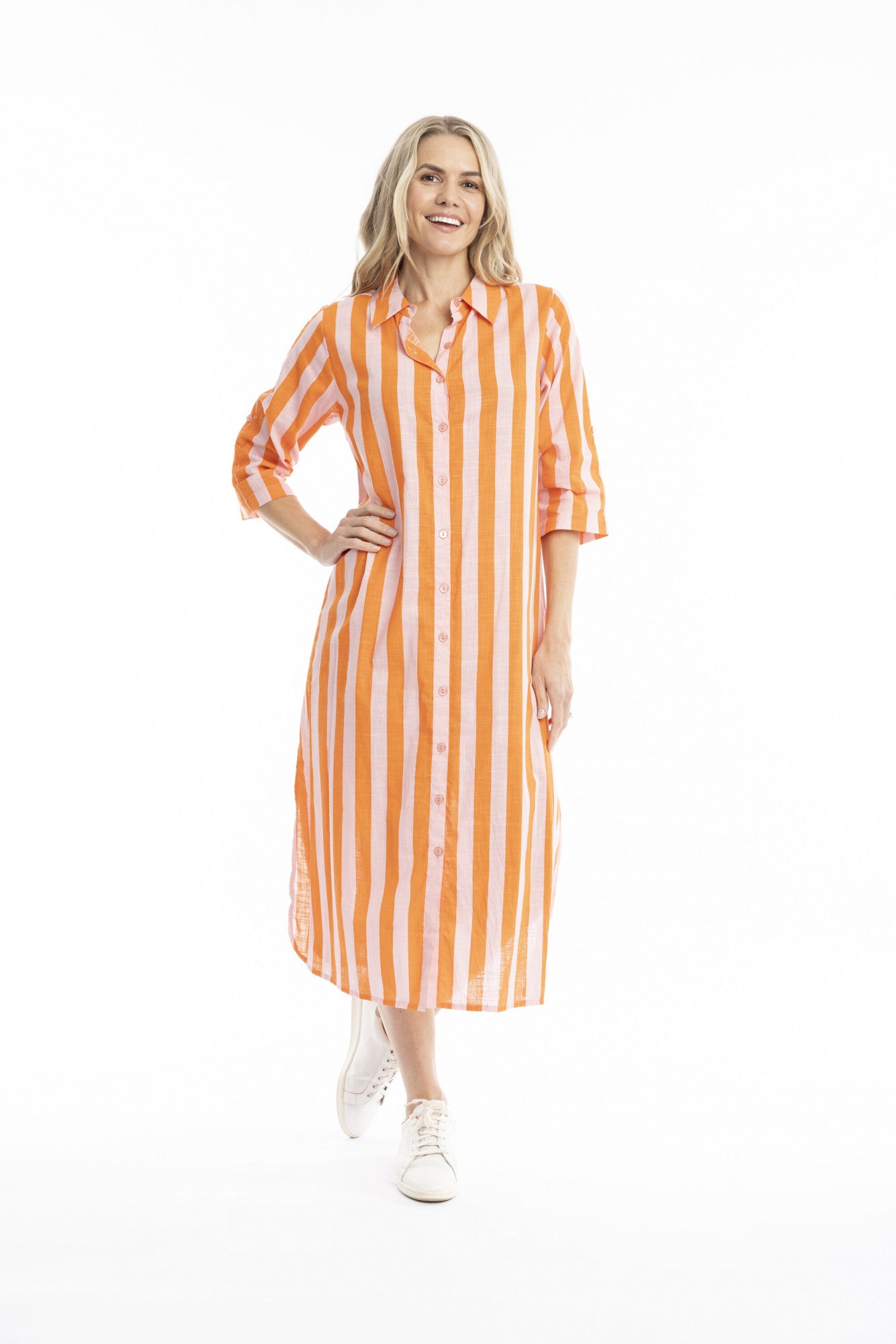 A woman modeling a long-sleeve midi shirt dress with vertical orange and white stripes, paired with white sneakers, against a plain white studio background.