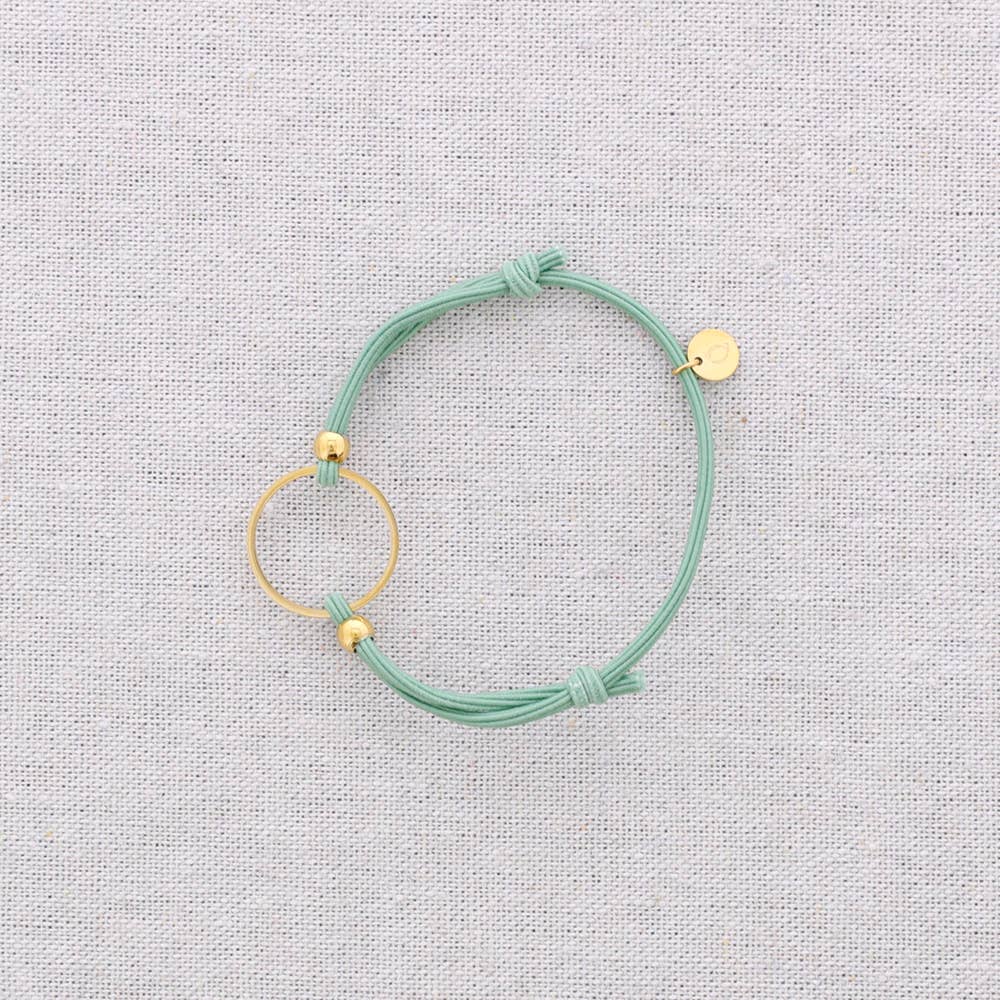 A mint green cord bracelet with gold-toned metal accents, including a circular ring, small beads, and a flat disc charm, displayed on a textured light gray fabric background.