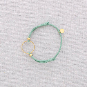 A mint green cord bracelet with gold-toned metal accents, including a circular ring, small beads, and a flat disc charm, displayed on a textured light gray fabric background.