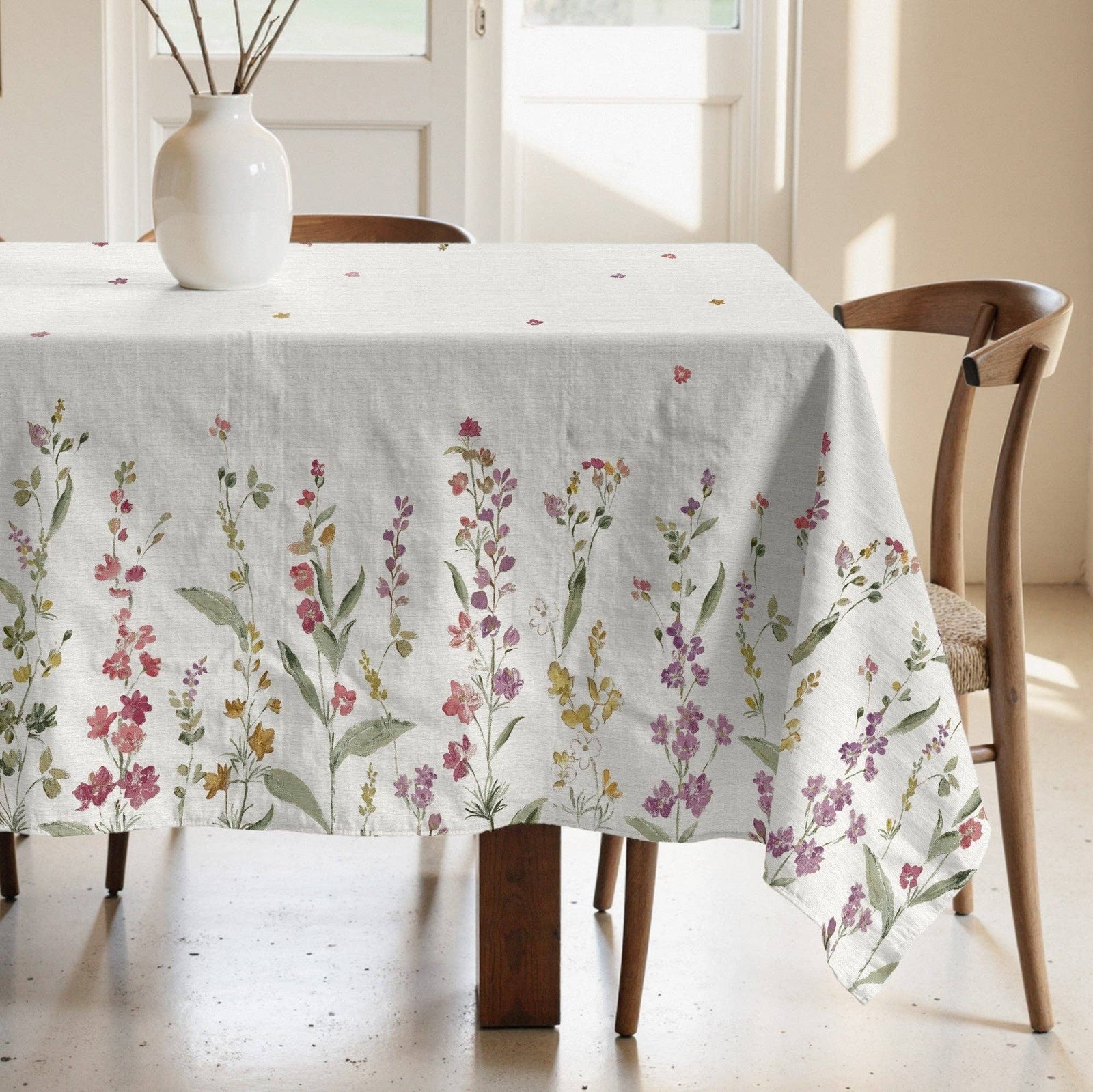 A white tablecloth with a delicate multicolor floral print draped over a wooden dining table in a bright, sunlit room.