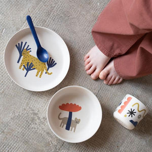 Children's tableware set with a plate, bowl, and spoon on a textured surface.