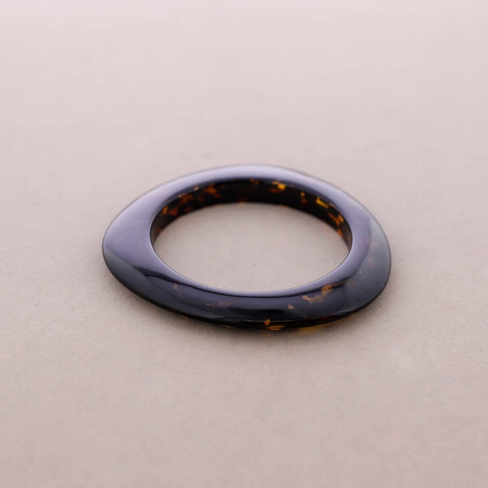 A single thick, rounded bangle with a glossy dark tortoiseshell pattern, in warm brown and amber mottled details, resting on a neutral beige background.