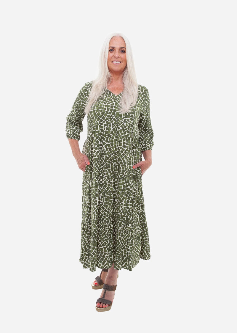 Woman wearing a blue patterned dress standing indoors with a wooden floor and white wall.