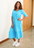 Woman wearing a blue patterned dress standing indoors with a wooden floor and white wall.