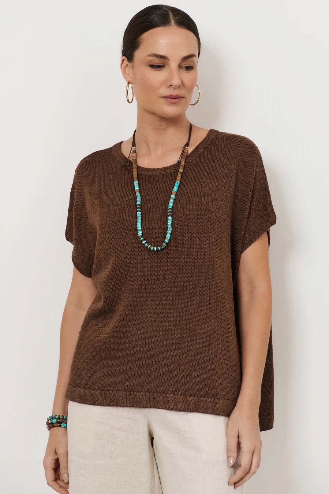 Woman wearing a brown top and beige pants with a white background with a turquoise and wood necklace