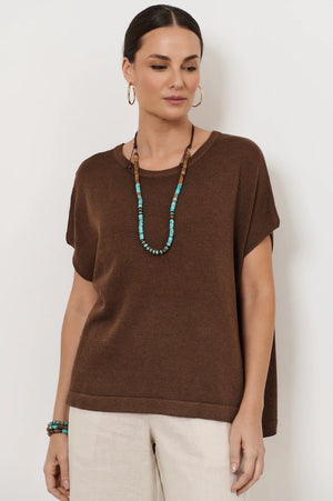 Woman wearing a brown top and beige pants with a white background with a turquoise and wood necklace