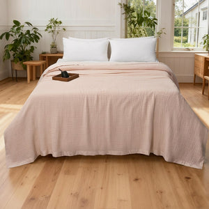 A bed dressed in a light blush pink textured waffle-weave bedding set, including a coverlet and two white pillows, styled in a bright, modern bedroom with light wood floors and plants.