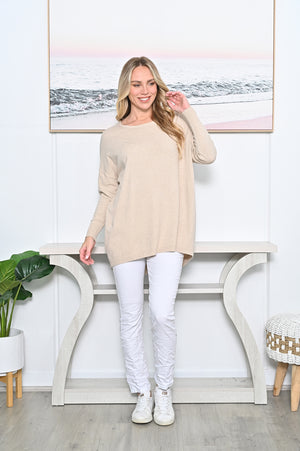 Woman wearing a beige sweater and white pants standing in a room with a decorative wall piece.