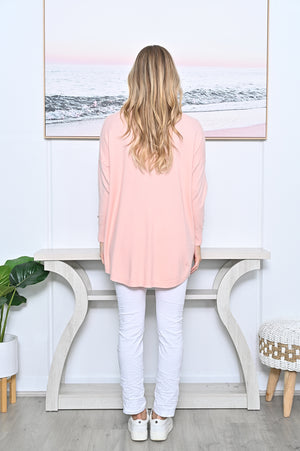 A woman with long blonde hair stands with her back to the camera wearing a light pink long-sleeve jumper, white pants, and white sneakers in a brightly lit room with a console table and framed wall art.