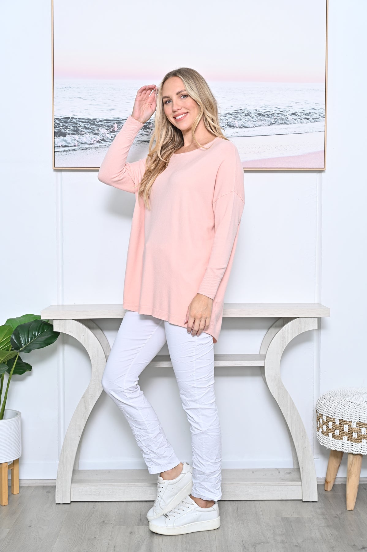 A woman modeling a light pink long-sleeve crewneck jumper, paired with white pants and white sneakers, standing in a brightly lit room with a console table and a framed picture on the wall.