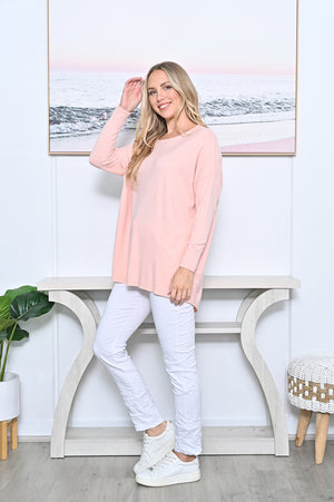 A woman modeling a light pink long-sleeve crewneck jumper, paired with white pants and white sneakers, standing in a brightly lit room with a console table and a framed picture on the wall.