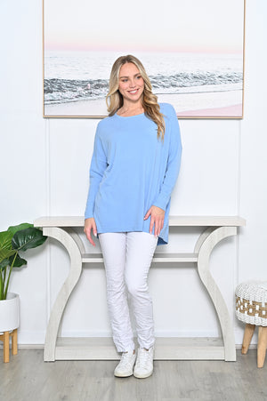 Woman wearing a light blue sweater and white pants standing in a room with a console table and plant.