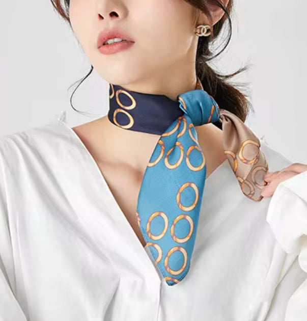 A woman wearing a blue silk scarf with a gold interlocking circles pattern, styled around her neck over a white blouse.