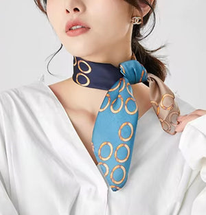 A woman wearing a blue silk scarf with a gold interlocking circles pattern, styled around her neck over a white blouse.