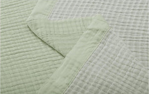 A close-up of a light sage green quilted cotton muslin blanket, showing its textured grid pattern and folded corner.