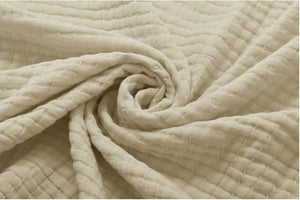 A close-up of a textured cream-colored fabric, likely cotton muslin or gauze, arranged in a spiral swirl to show its soft, quilted weave pattern.