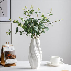 A white ribbed ceramic vase with a wavy, fluted design holding green eucalyptus branches with small pale flowers, styled on a white table next to a small wooden calendar and a white cup and saucer against a light gray wall.