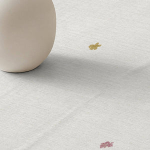 A close-up of a white textured fabric surface with a small gold flower and a small pink flower printed on it, alongside the lower edge of a matte beige ceramic vase.