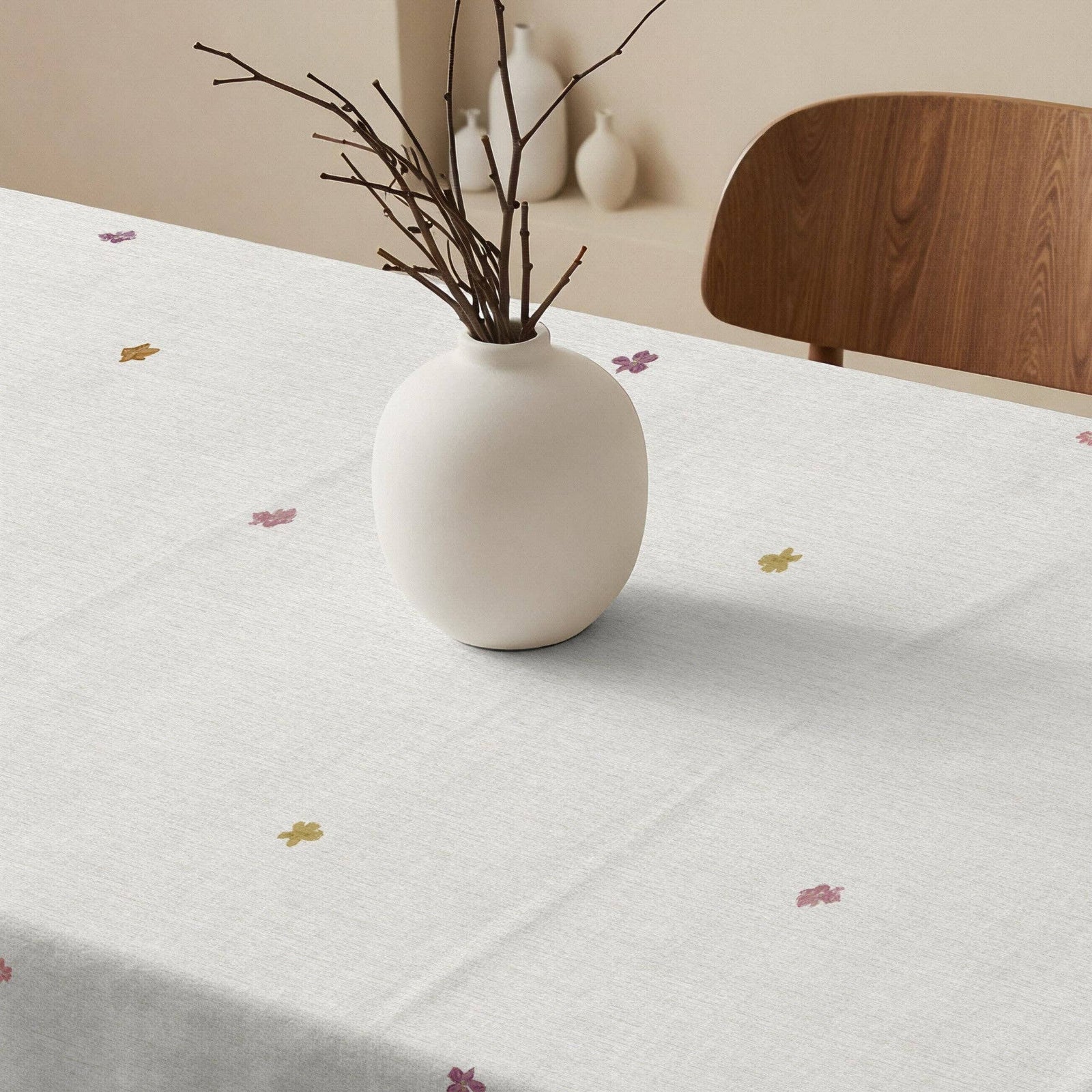 A white tablecloth with a delicate multicolor floral print draped over a wooden dining table in a bright, sunlit room.