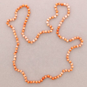 A beaded necklace arranged in an irregular outline on a plain light gray background. The beads are primarily orange with white marbled or speckled sections throughout.