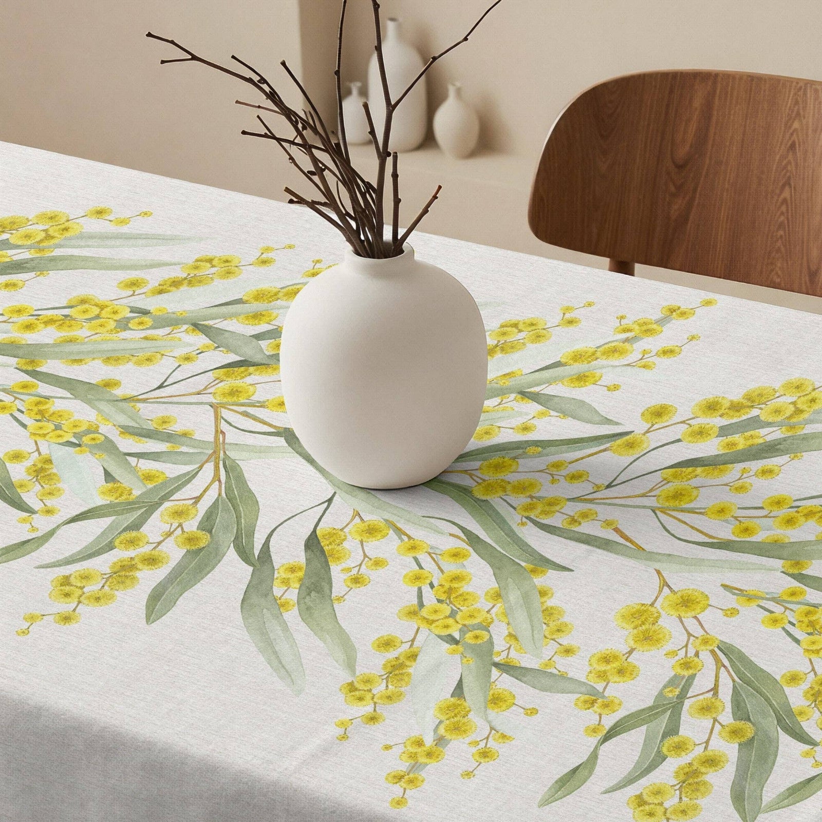 A white tablecloth with a yellow wattle blossom and green eucalyptus leaf print, styled on a dining table with a white ceramic vase containing bare branches and a wooden chair in the background.