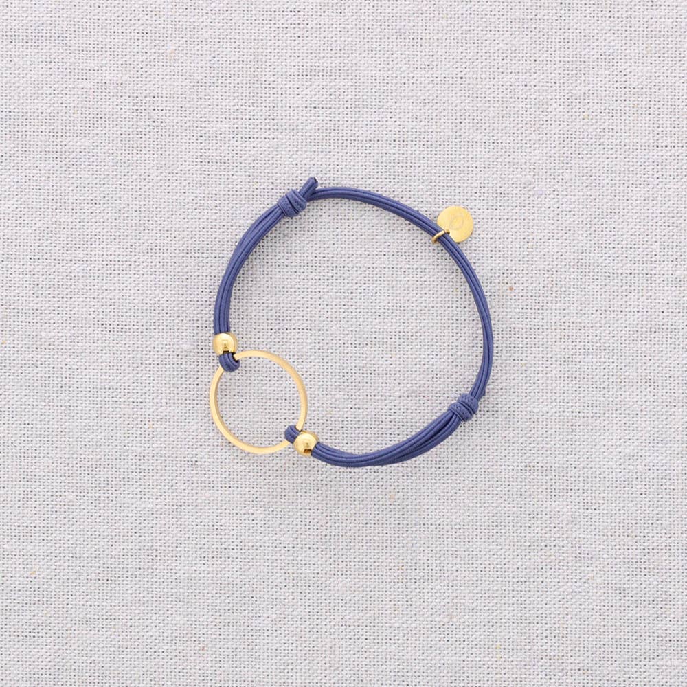 A mint green cord bracelet with gold-toned metal accents, including a circular ring, small beads, and a flat disc charm, displayed on a textured light gray fabric background.