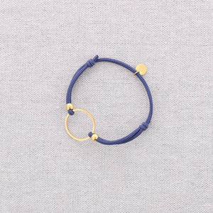 A Steel blue cord bracelet with gold-toned metal accents, including a circular ring and small beads, displayed on a textured light gray fabric background.