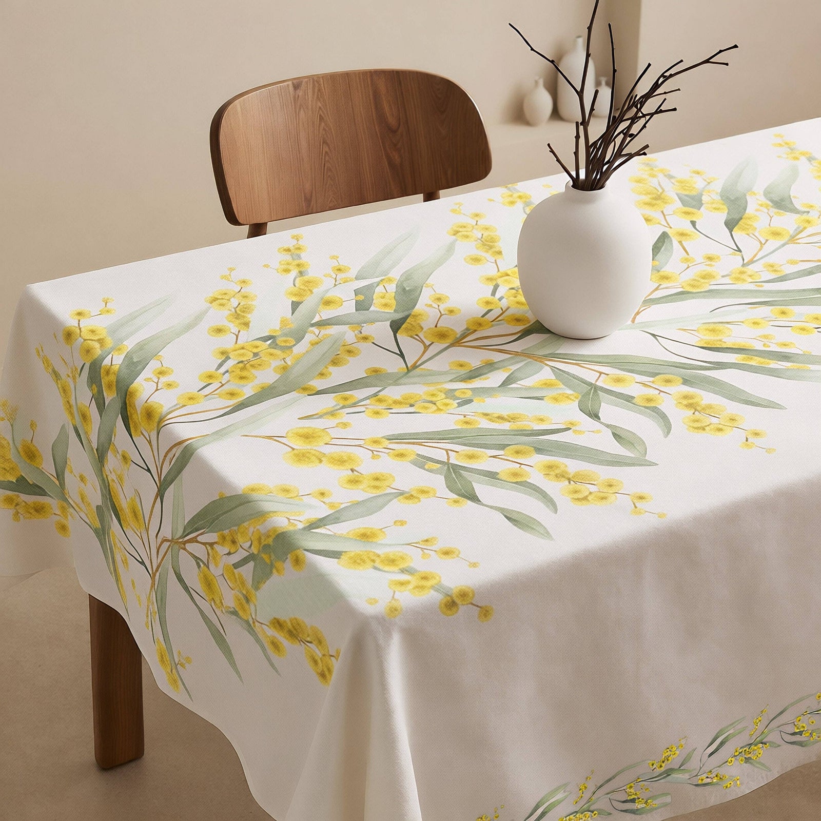 A white tablecloth with a yellow wattle blossom and green eucalyptus leaf print, styled on a dining table with a white ceramic vase containing bare branches and a wooden chair in the background.
