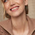 A woman wearing gold earrings with a green gemstone against a neutral background
