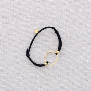 A black elastic cord bracelet with a gold-tone circular ring and small gold disc bead, displayed on a textured light gray fabric background.