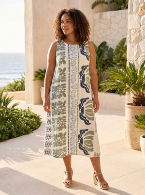 A woman wearing a sleeveless midi dress with a botanical leaf pattern in green, navy, and blue on an off-white background, standing on a sunlit patio with plants and the ocean in the background.