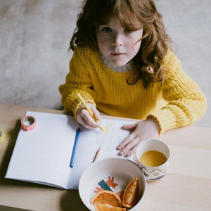 Child in a yellow sweater drawing at a table with a bowl and cup.