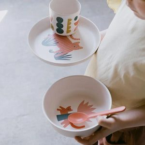Child holding a bowl with a spoon, next to a plate and cup with animal designs on a light background