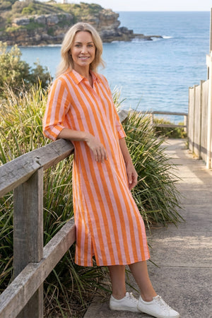 A woman wearing a bright orange and white vertically striped midi shirt dress stands on a coastal pathway next to a wooden railing, with the ocean and cliffs in the background.