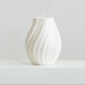 A white ceramic vase with a textured, vertically wavy ribbed design, displayed on a white surface against a light gray background.